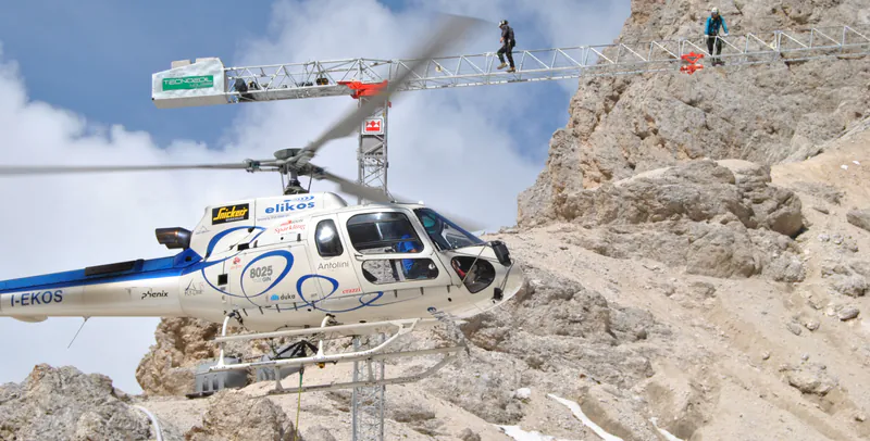 A helicopter beside a Tecnoedil crane at a mountain construction site
